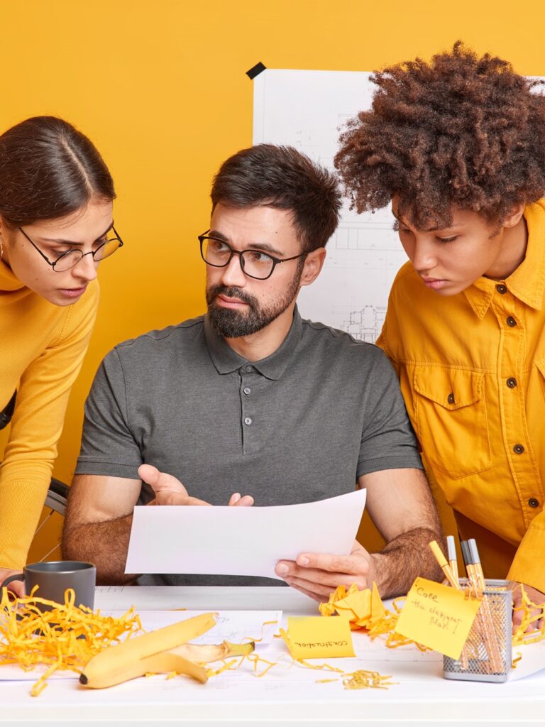 Group of international architects discuss ideas for engineering project enjoy working process together pose at desktop concentrated at paper yellow background. Diverse colleagues inn office.